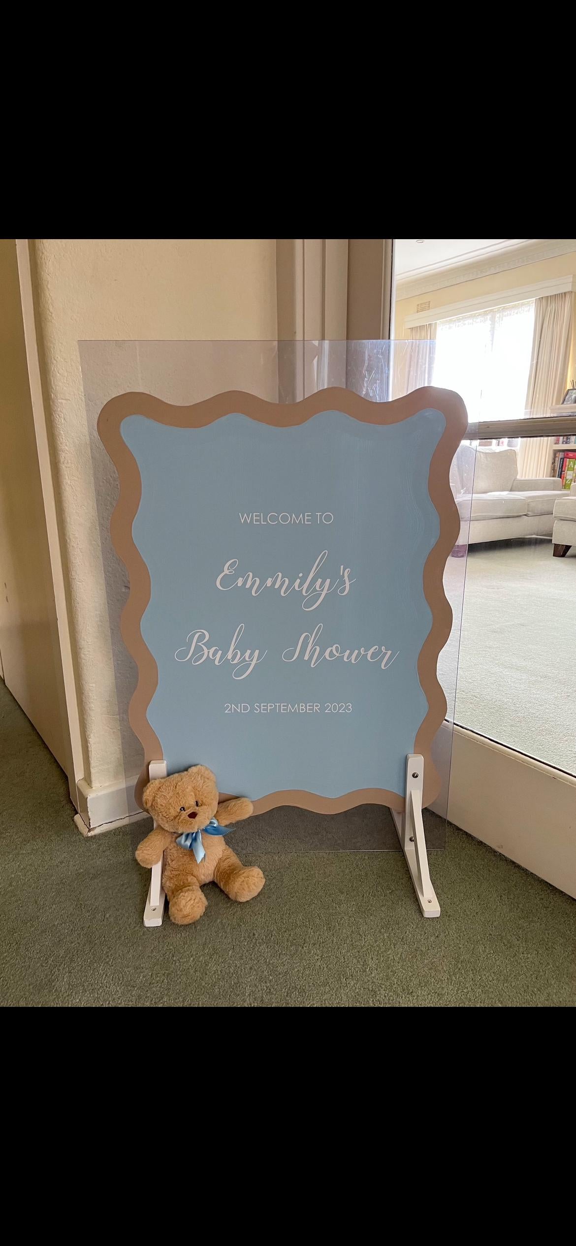 Baby shower sign with teddy bear on a stand in a room.