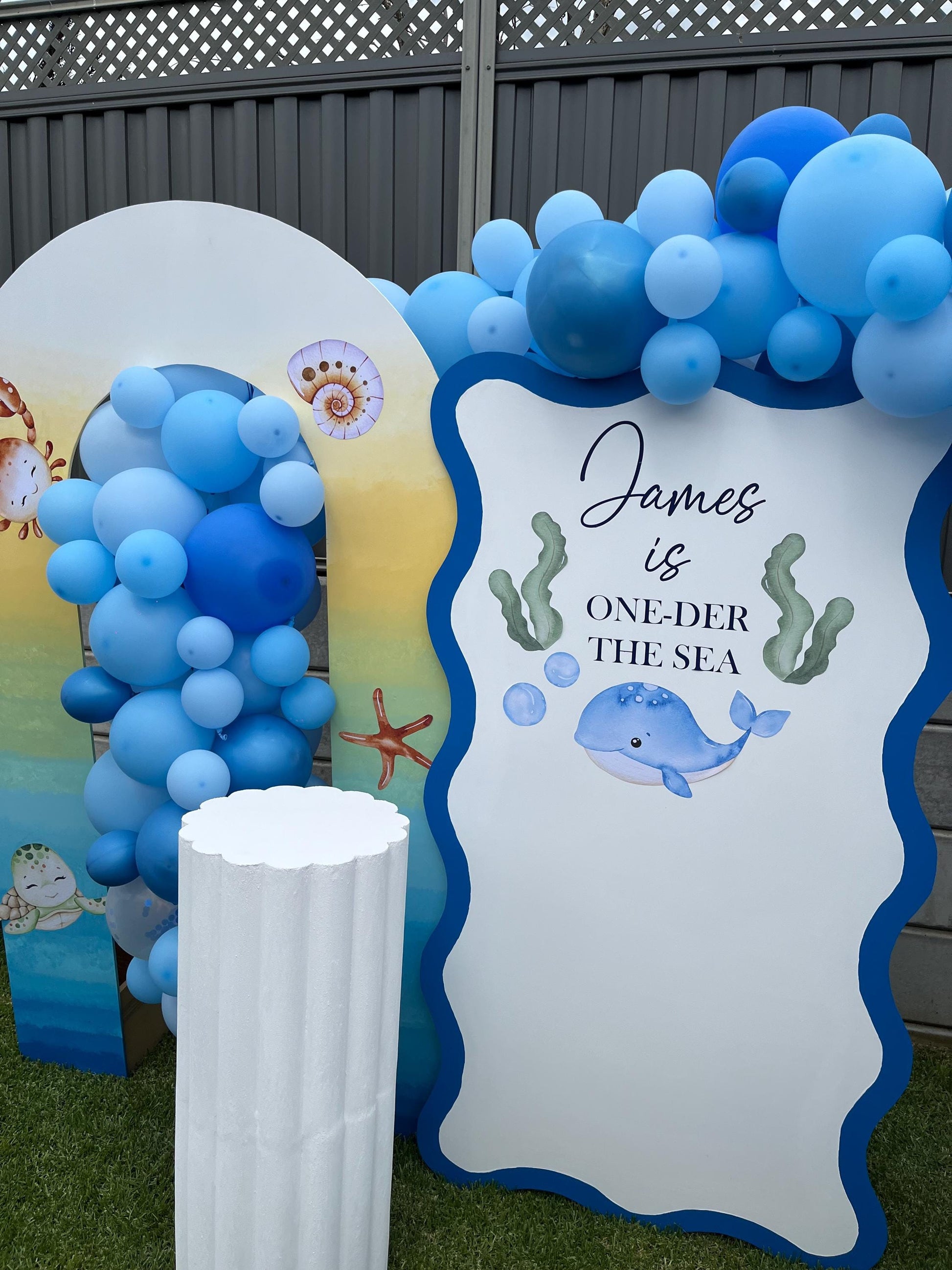 Decorative setup with blue balloons and a sign reading 'James is ONE-der the Sea' against a fence.