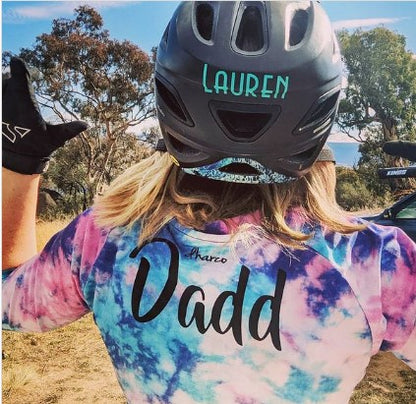Person wearing a tie-dye shirt with 'Dadd' and a helmet with 'LAUREN' on it, outdoors.